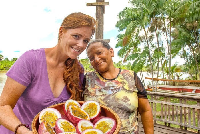 Two people smile while holding a bowl filled with halved passion fruits outdoors. The background features palm trees and a wooden structure beside a river, conveying a tropical atmosphere.