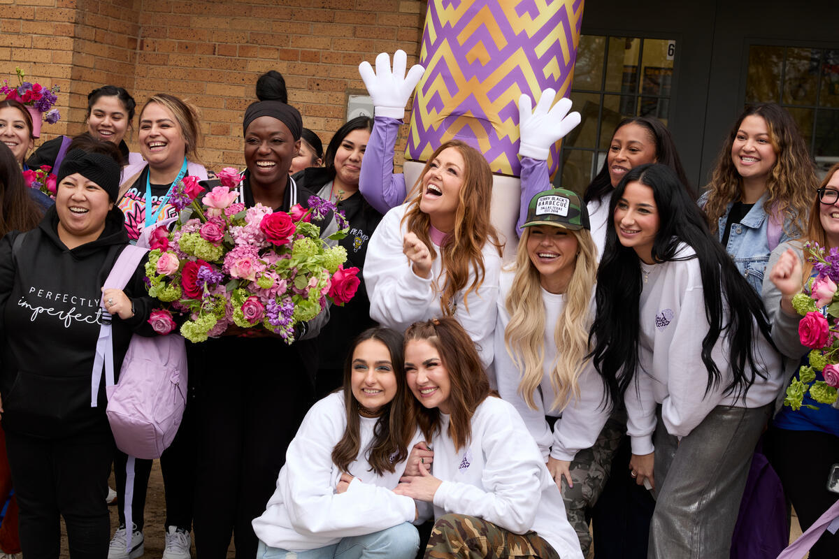 A group of smiling people, some holding flowers, gather in front of a brick building. A person wears a hoodie that reads Perfectly imperfect. A large inflatable object with purple patterns is present.