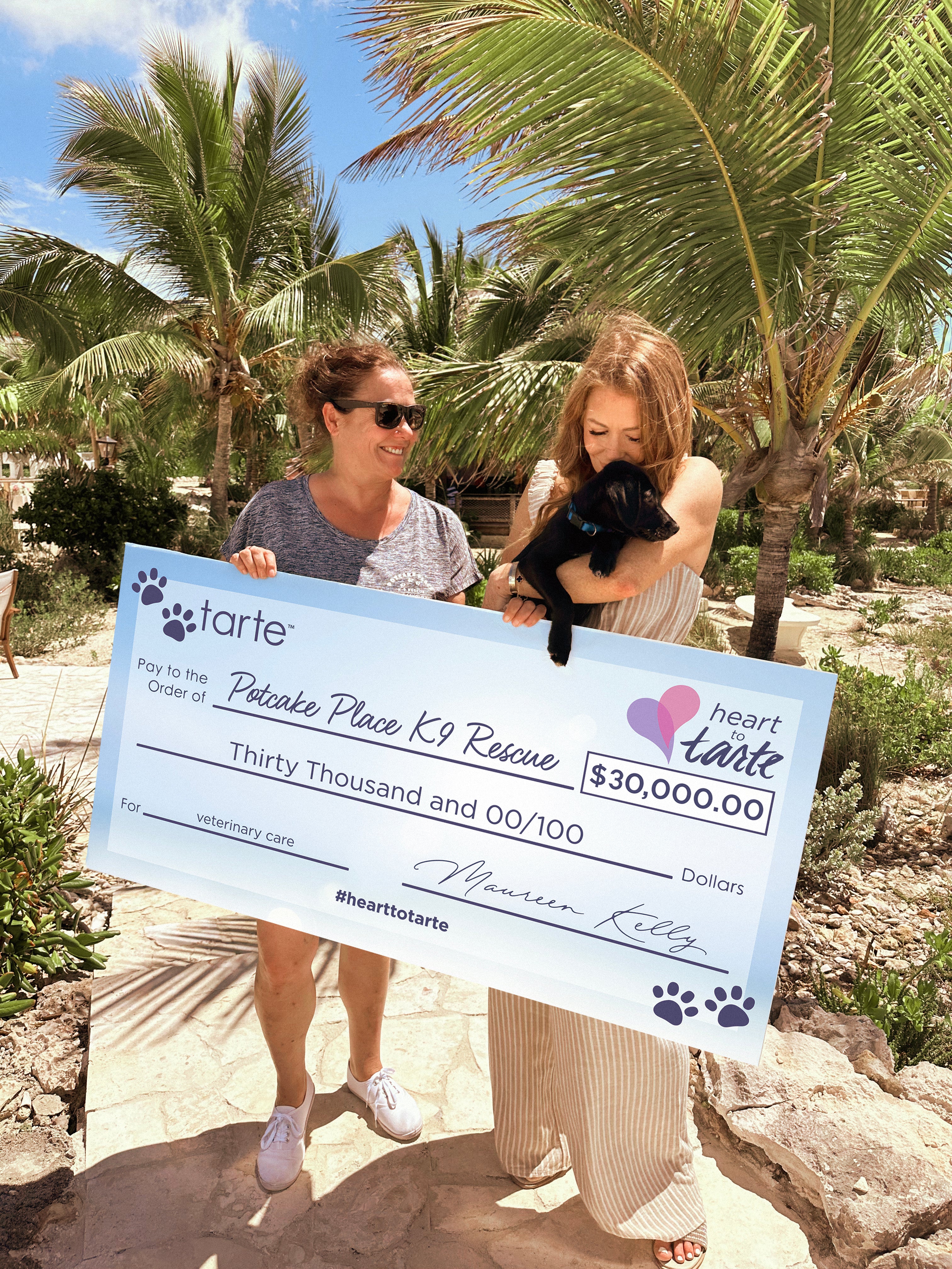 Two people hold a large check under palm trees. One holds a black puppy. The check reads: Tarte Cosmetics. Pay to the Order of Potcake Place K9 Rescue, Thirty Thousand Dollars, hearttotarte.